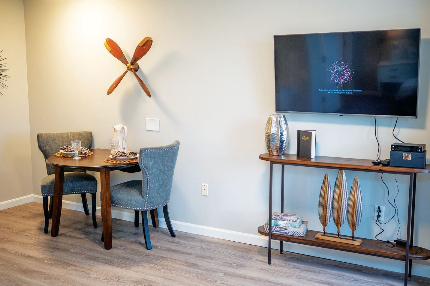 A modern living room features a small wooden dining table with two grey chairs, a wall propeller, and a wall-mounted TV.