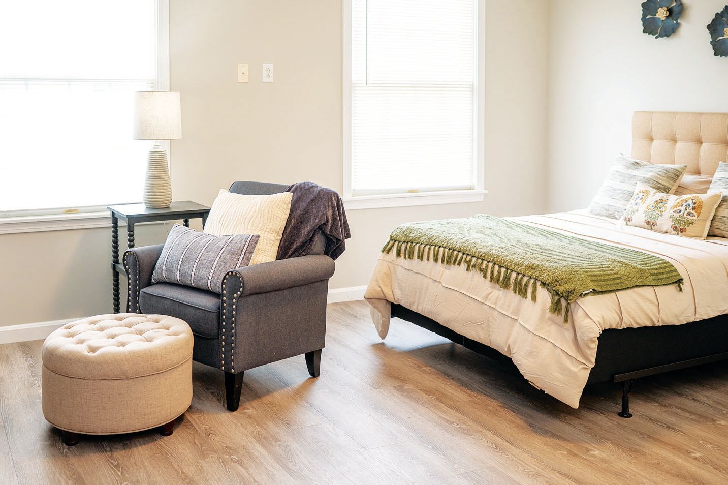 A bedroom with a gray armchair, a tan tufted ottoman, a side table with a lamp, and a bed with neutral-toned bedding.