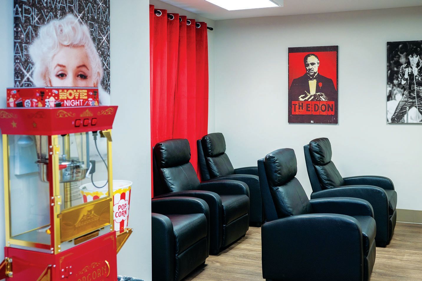 A home theater room with three black recliner chairs, a red popcorn machine, and movie-themed wall posters.