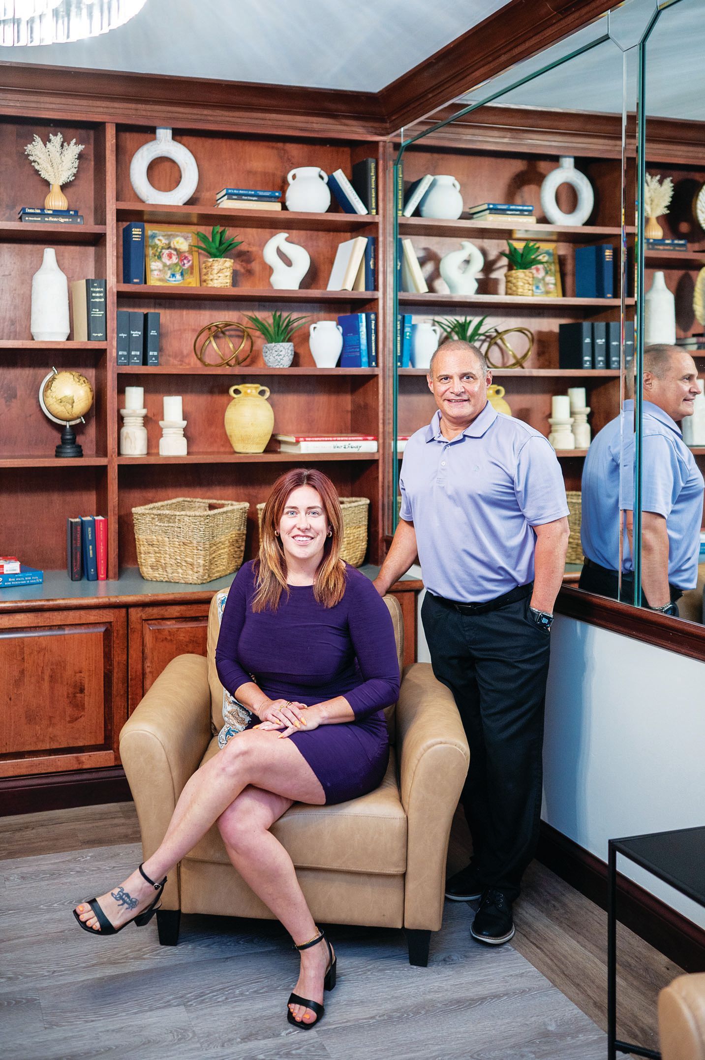 A smiling woman in a purple dress sits in a tan chair next to a man in a blue polo, posed before a wooden bookshelf.