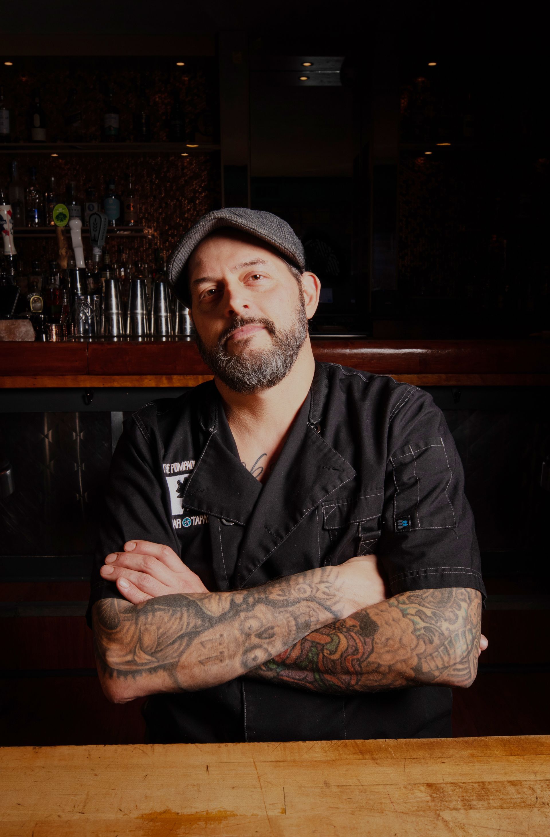 A person with tattoos on their arms stands with arms crossed, wearing a black chef coat and cap in a dimly lit bar setting.