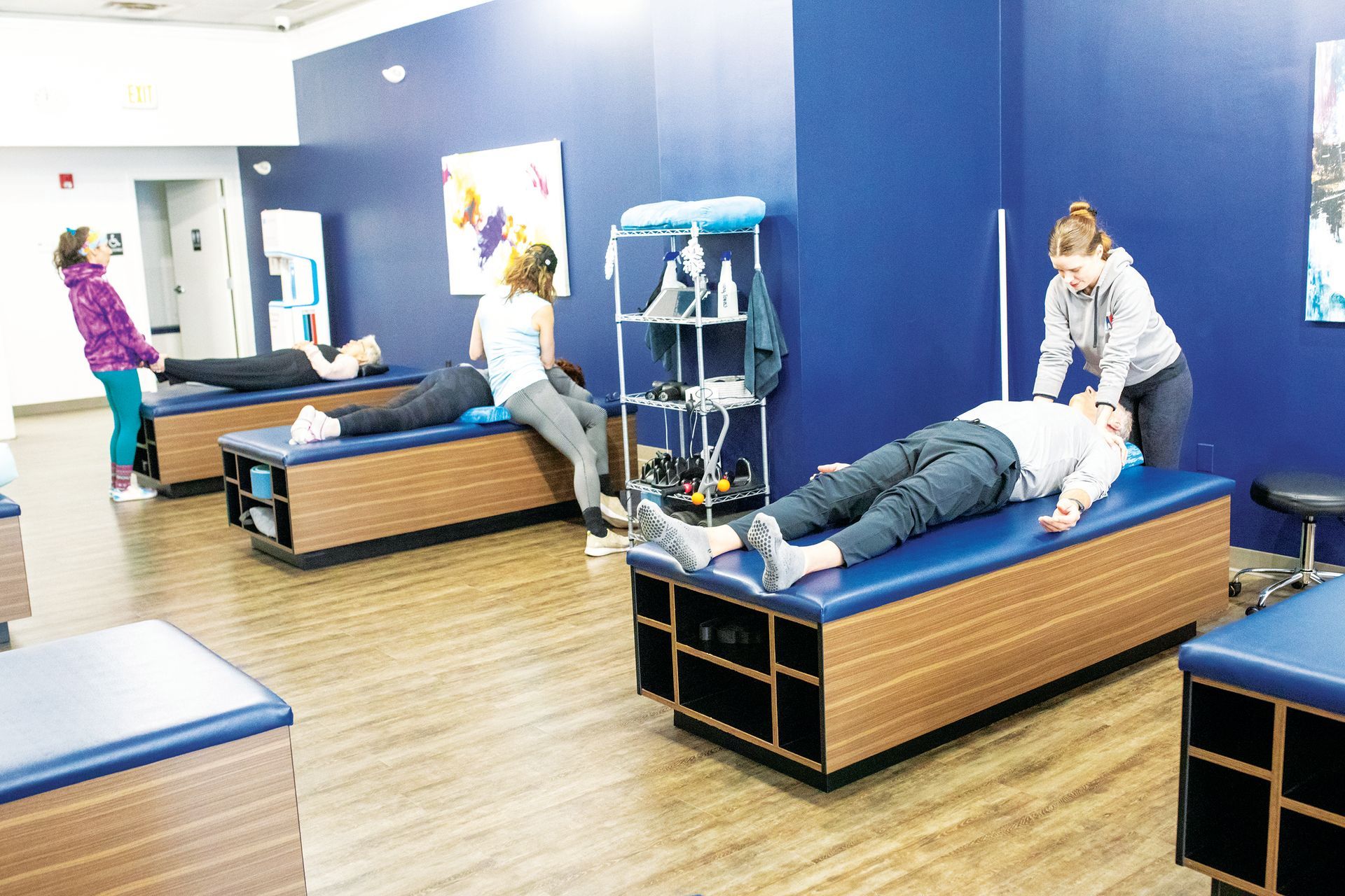 A physical therapy training lab with students practicing hands-on patient care on treatment tables in a blue-walled room.