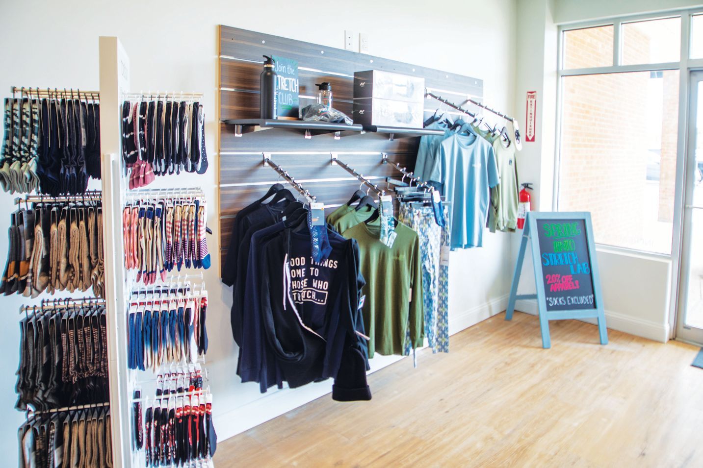 Clothing store retail display with racks of apparel and accessories, a wooden floor, and a small standing chalkboard sign.