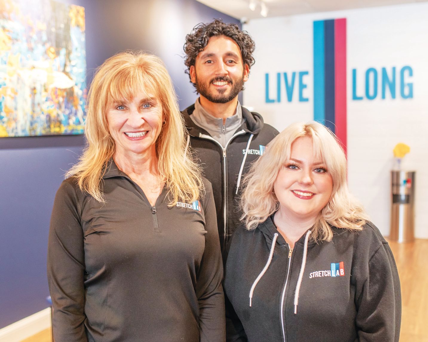 Three people wearing branded clothing stand smiling in an office with a wall sign reading 