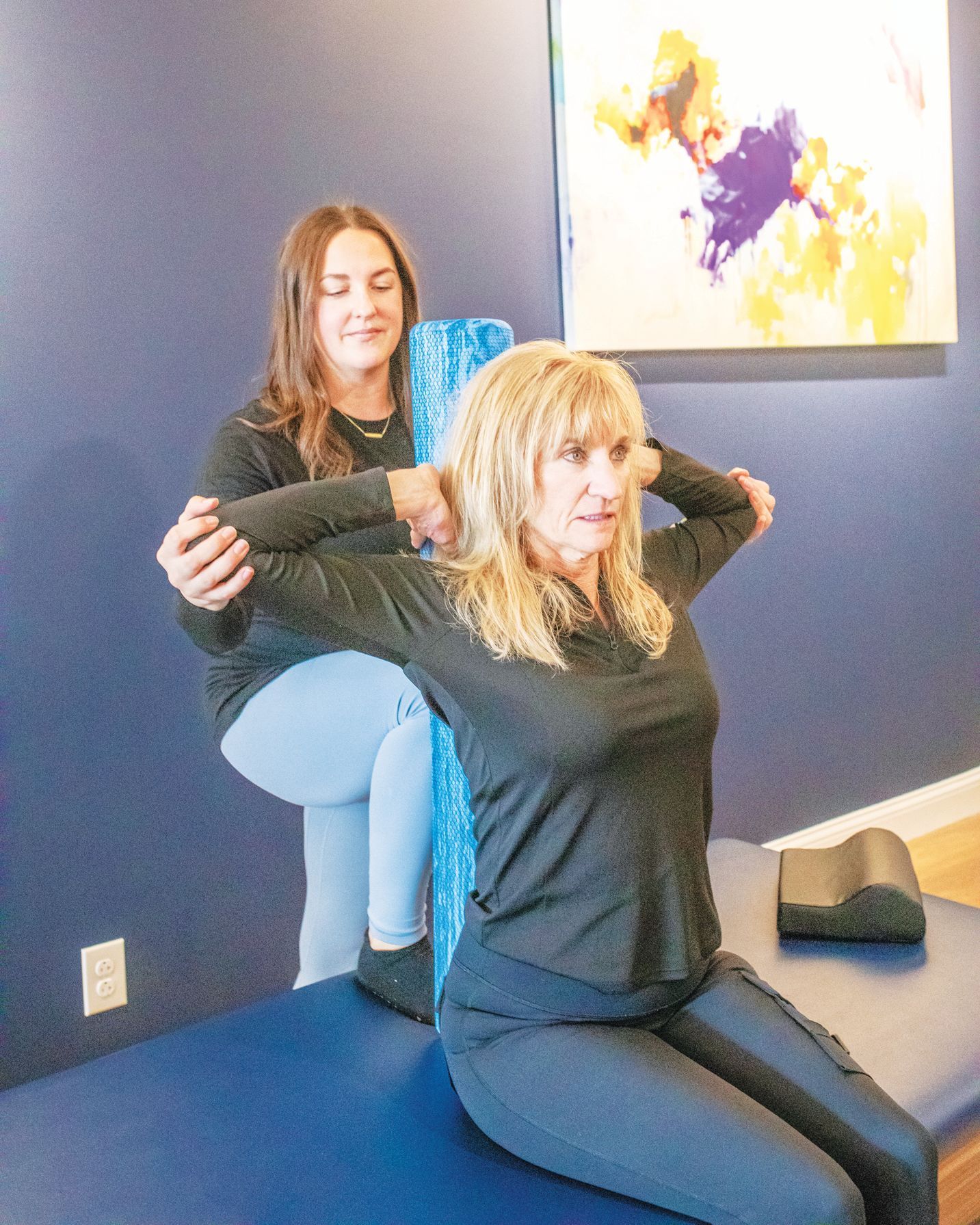 A person in dark clothes assists another with a foam roller stretch, placing their hands on their upper arms while seated.