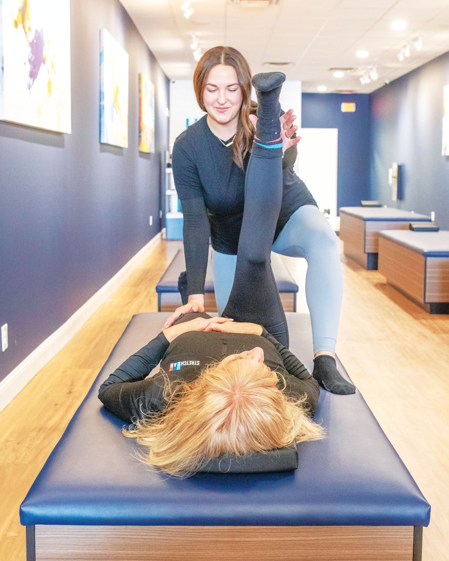 A practitioner stretches the raised leg of a client lying on a blue treatment table in a wellness studio.