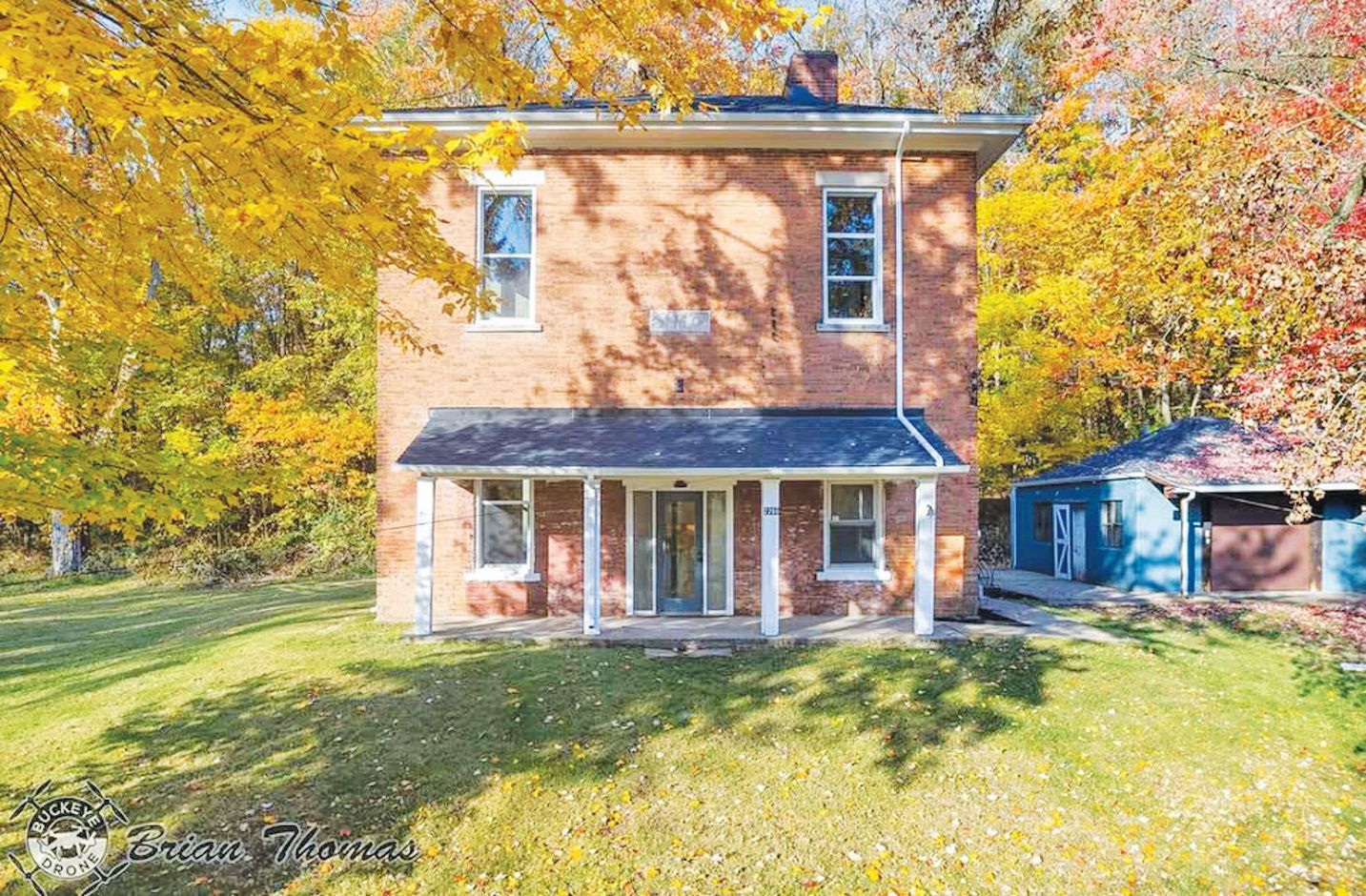A two-story red brick house with a covered front porch, surrounded by trees with yellow and autumn foliage.