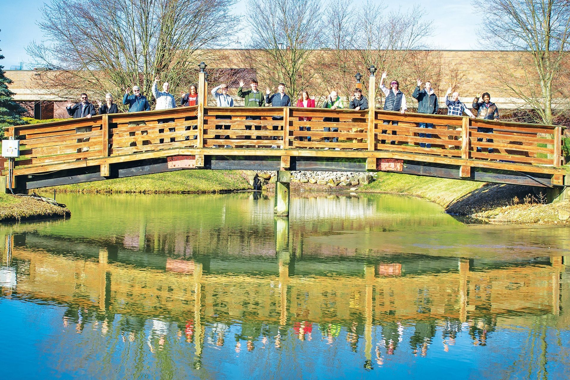 A group of people standing on a wooden footbridge over a pond, waving at the camera on a sunny day.