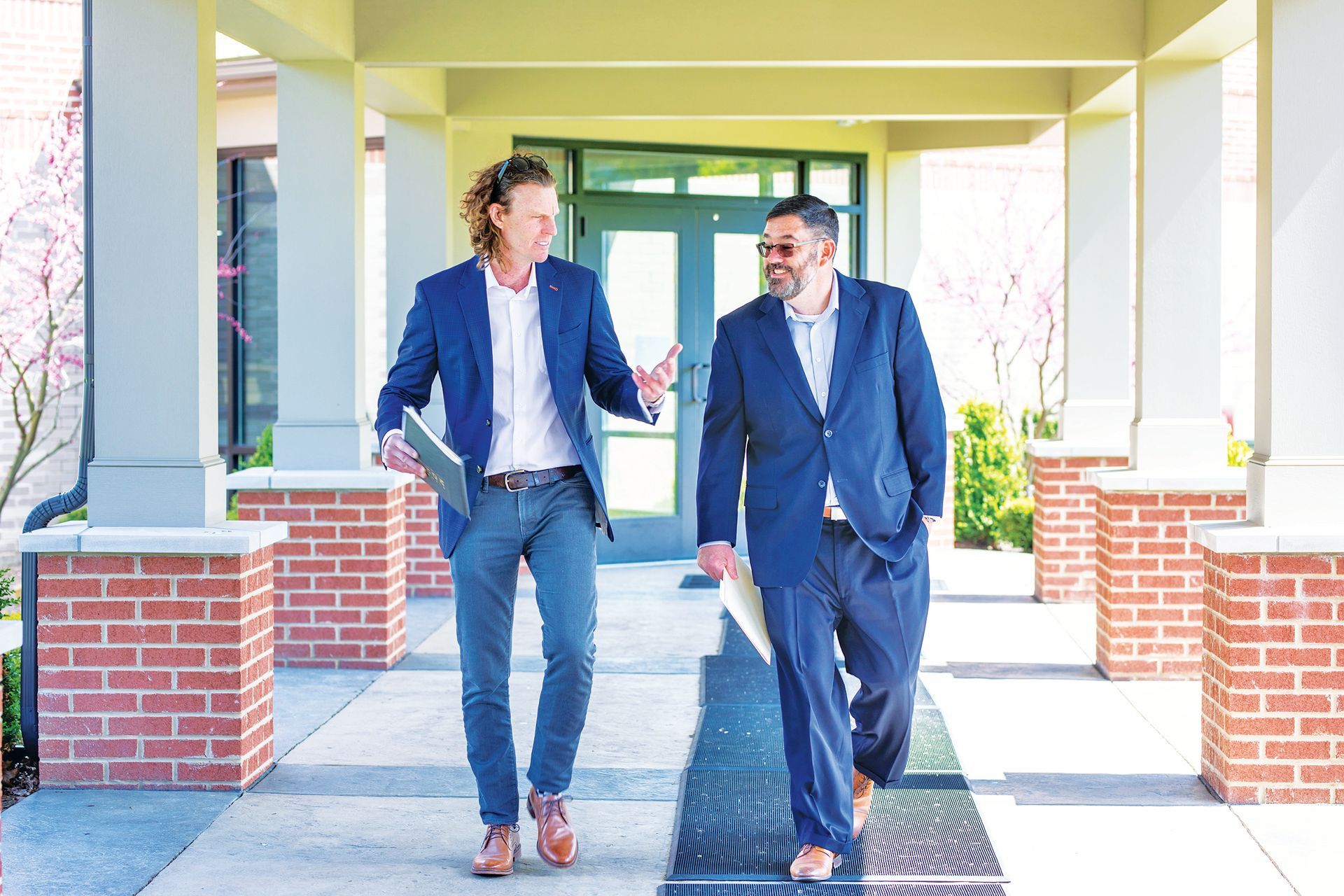 Two people in professional attire talk and walk together on a pathway outside a building with brick columns.