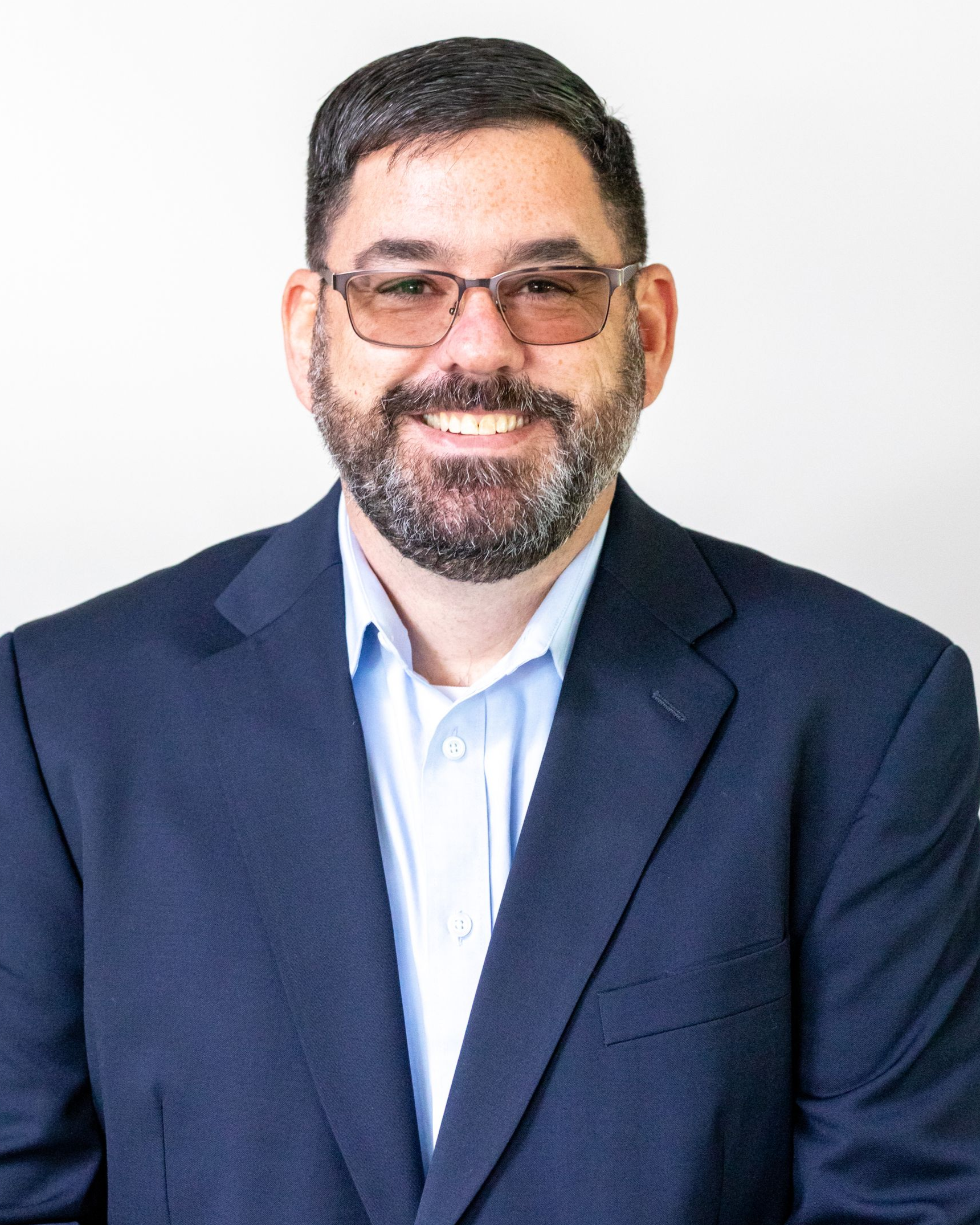 A smiling person with a beard and glasses wears a light blue dress shirt and a navy blue blazer against a white background.