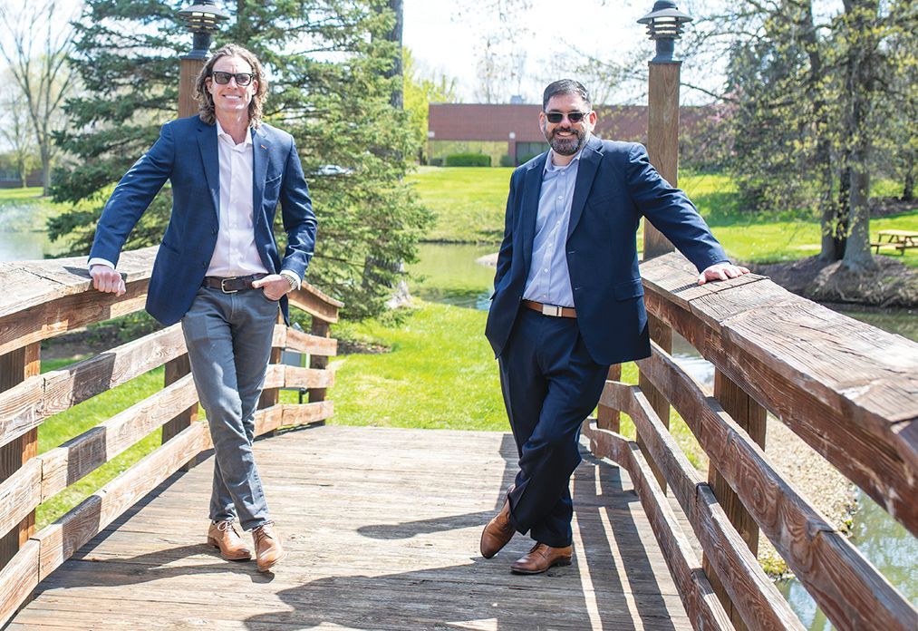 Two men in blazers and dress slacks pose on a wooden bridge in a grassy park setting.