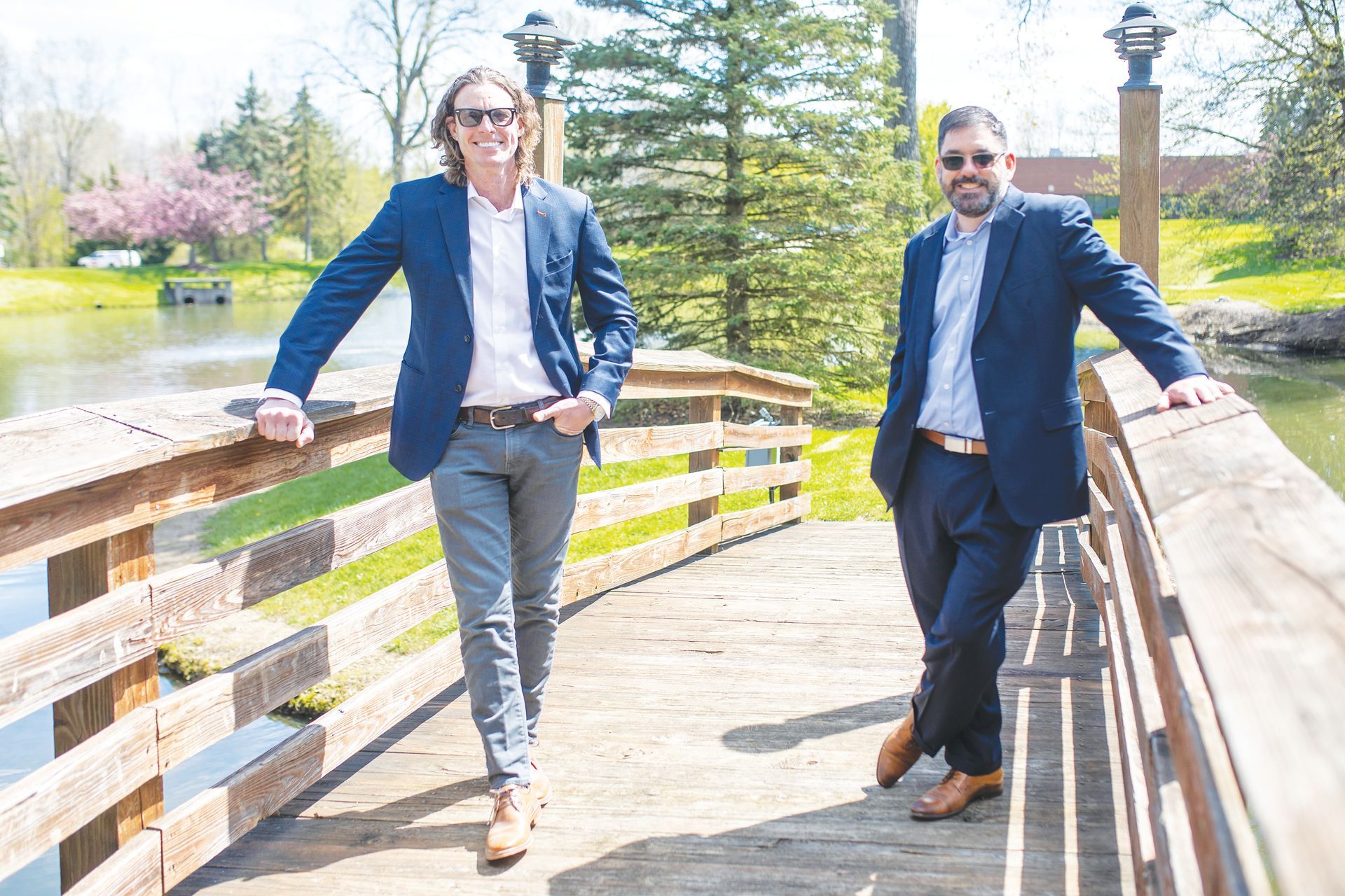 Two professionals in blue blazers standing on a wooden bridge over a pond in a park on a sunny day.