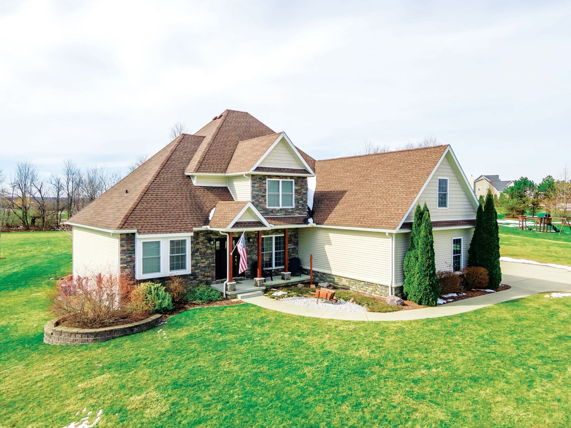 A two-story suburban house with brown roofing, beige siding, and stone accents, set on a grassy lawn under a clear sky.