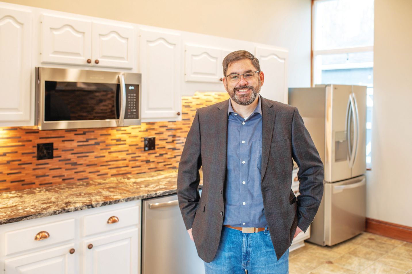 A smiling person in a blazer and jeans stands in a kitchen with white cabinets, stainless appliances, and a tiled backsplash.