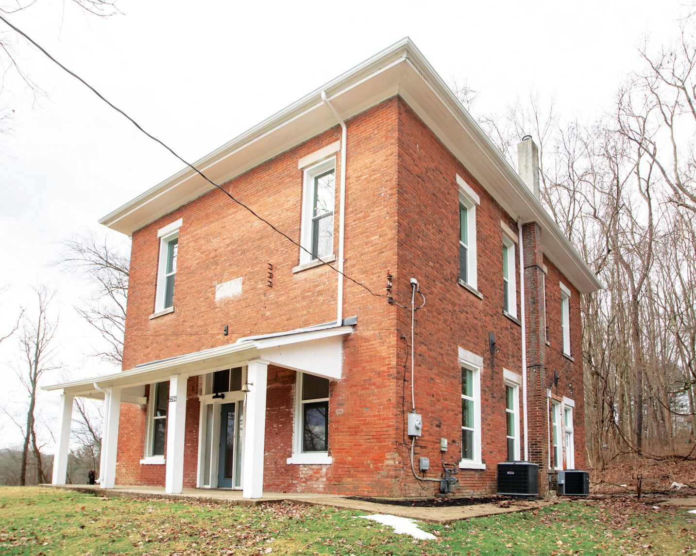 A two-story red brick house with a white front porch and overhanging roof sits on a grassy lot surrounded by bare trees.