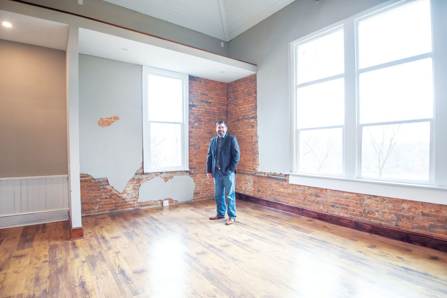 A person stands in a sunlit room featuring hardwood floors, exposed brick walls, and tall windows.