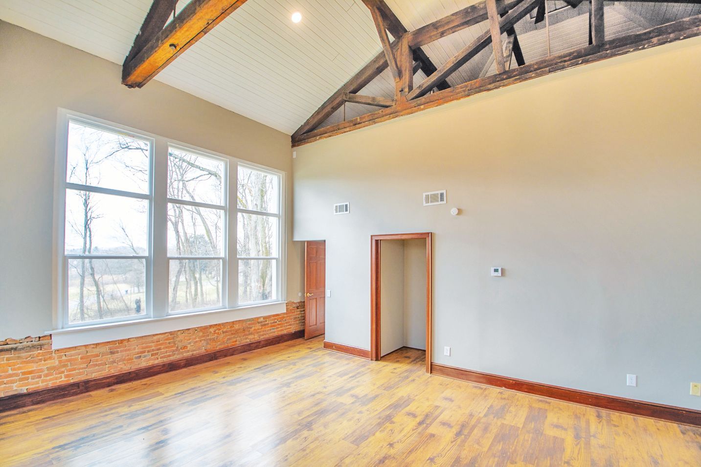 Spacious room with wood floors, exposed ceiling beams, large windows, and a doorway, set against light grey walls.
