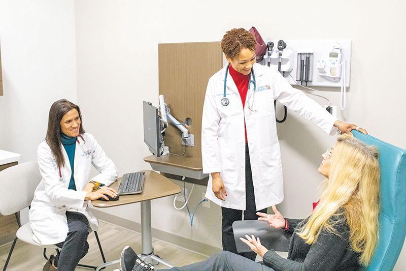 Two healthcare providers in white coats consult with a patient sitting in a clinical exam room.