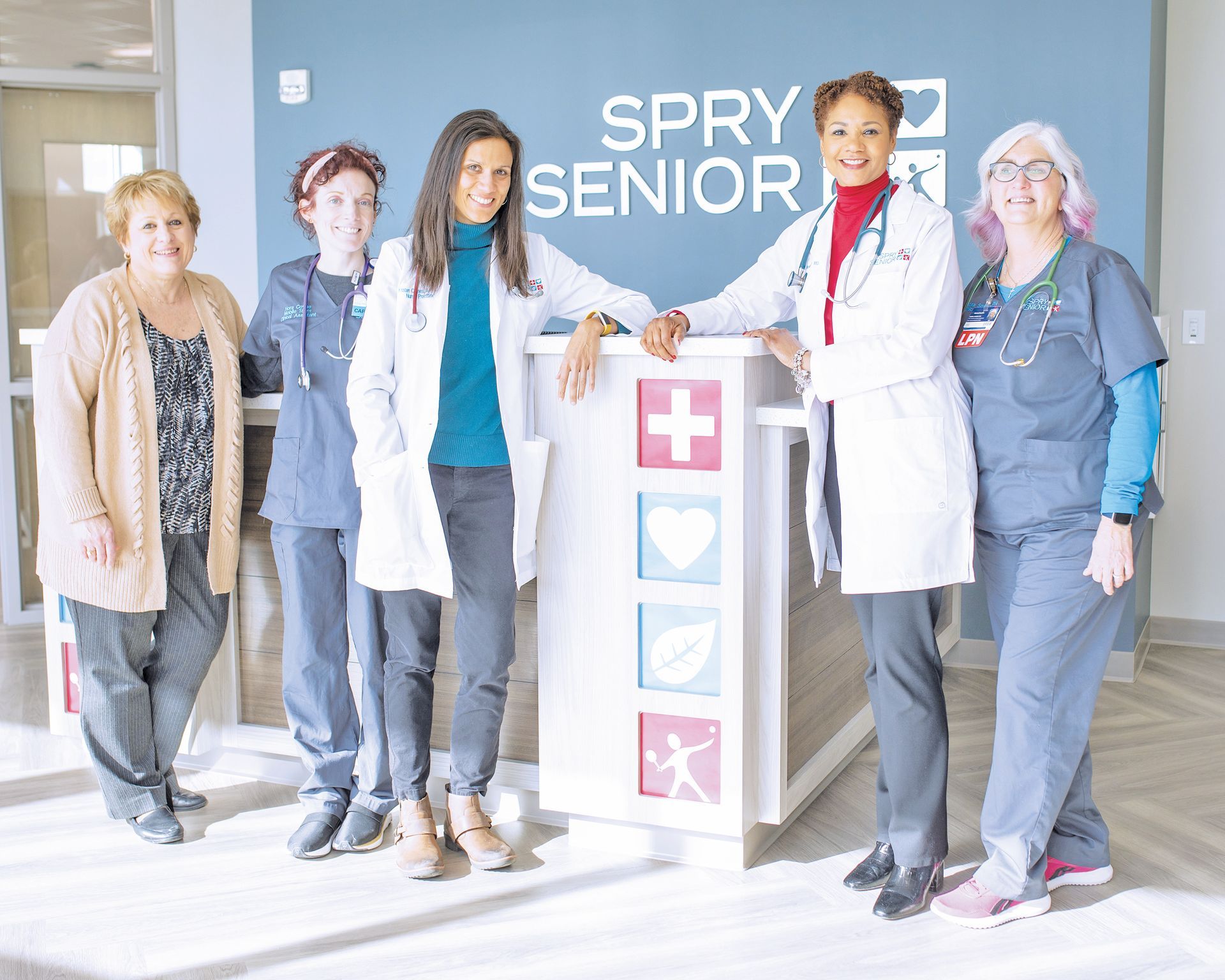 Five staff members pose together behind the front desk at Spry Senior, a medical facility with a logo on the back wall.
