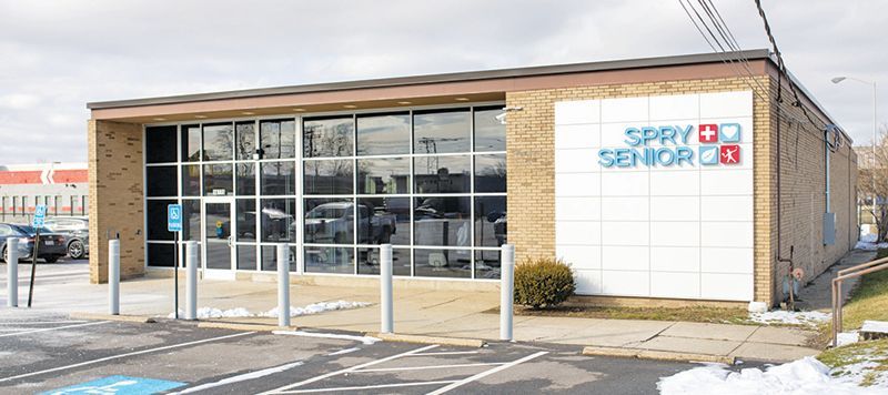 The Spry Senior center building with brick exterior, large glass windows, and a white sign with blue and red logos.