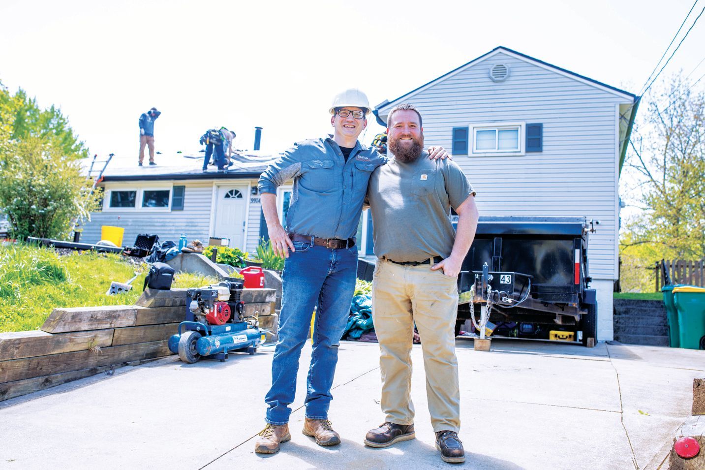 Two men smile while standing in front of a house under renovation with workers visible on the roof.