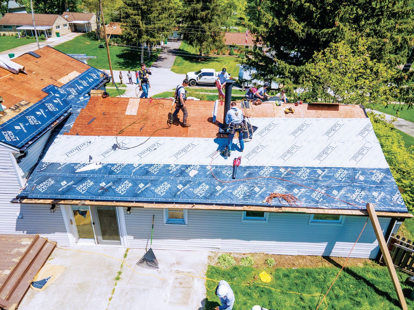 Roofers work on a house roof, installing layers of underlayment over the plywood deck with tools and materials nearby.