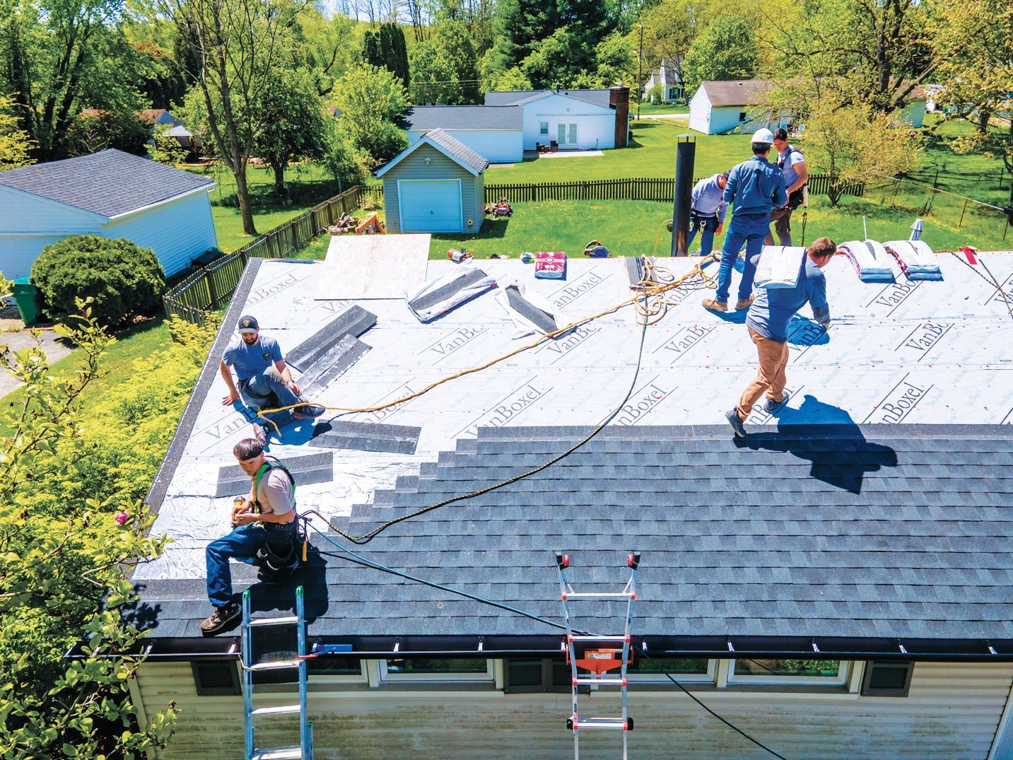 Four workers install dark shingles on a residential roof, with some wearing safety harnesses connected to ropes.