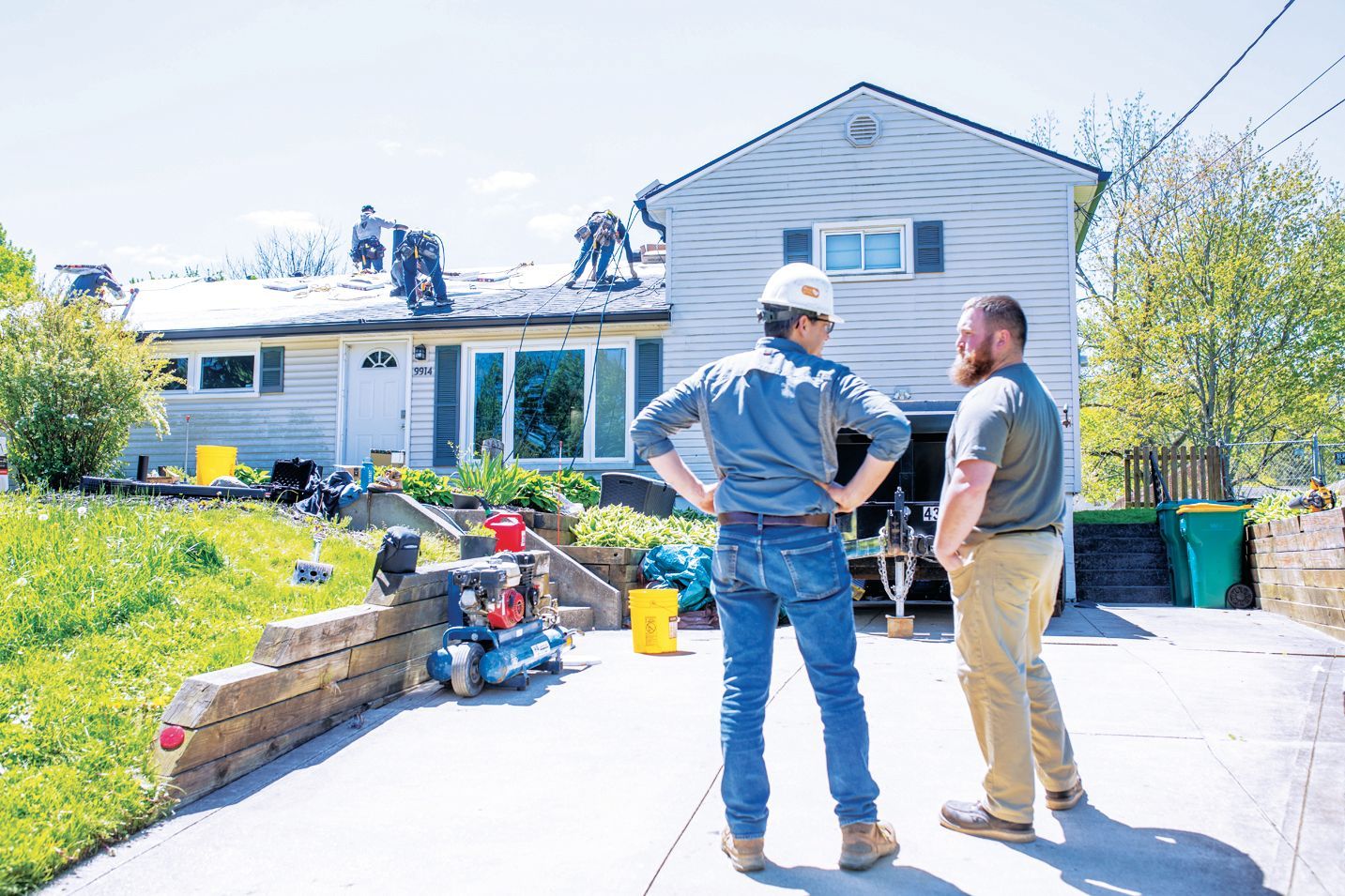 Two workers stand in a driveway talking, while others perform roofing repairs on a house in the background.
