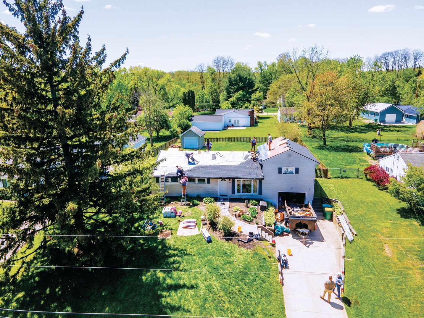 An aerial view shows workers installing shingles on a residential home roof on a sunny day with trees in the background.