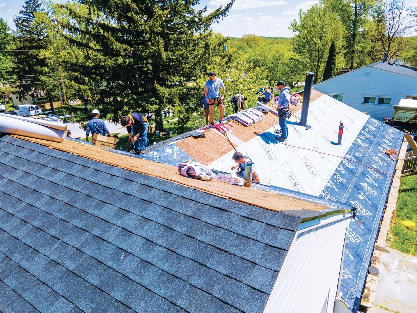 Workers in casual clothing install dark blue shingles on a house roof under a sunny sky surrounded by trees.