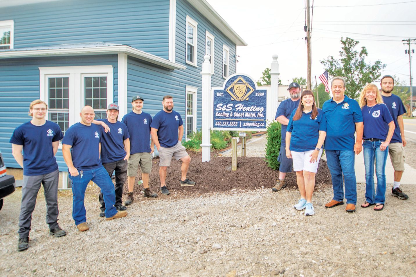 A group of people in blue shirts stand outside a business office in front of a sign that reads S&S Heating and Cooling.