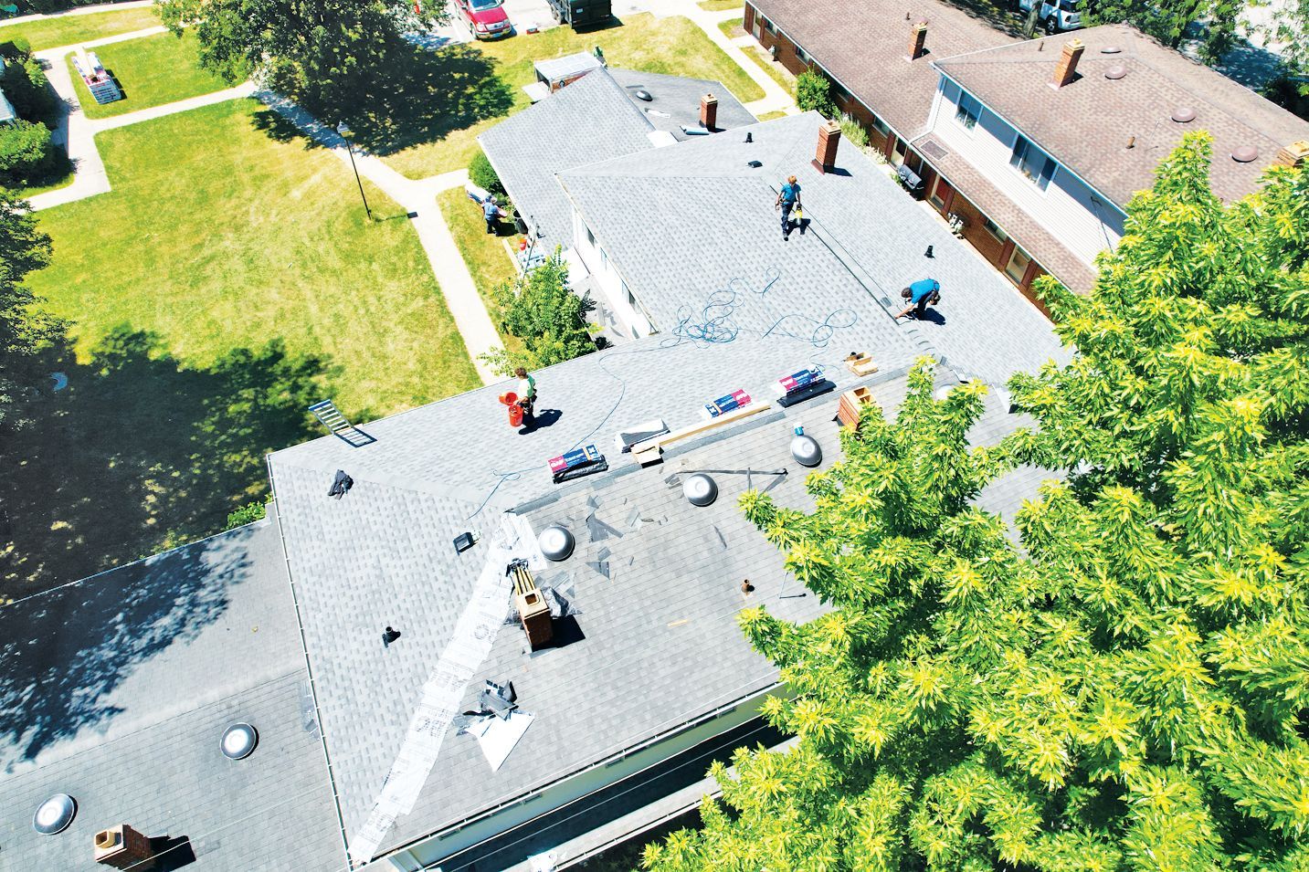 An aerial view shows workers repairing a large, shingled roof on a sunny day with trees and lawn nearby.