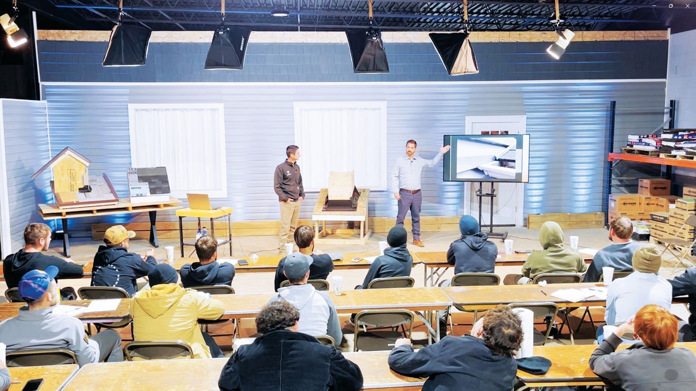 Two instructors lead a training session for an audience seated in a room, with one pointing to a presentation screen.