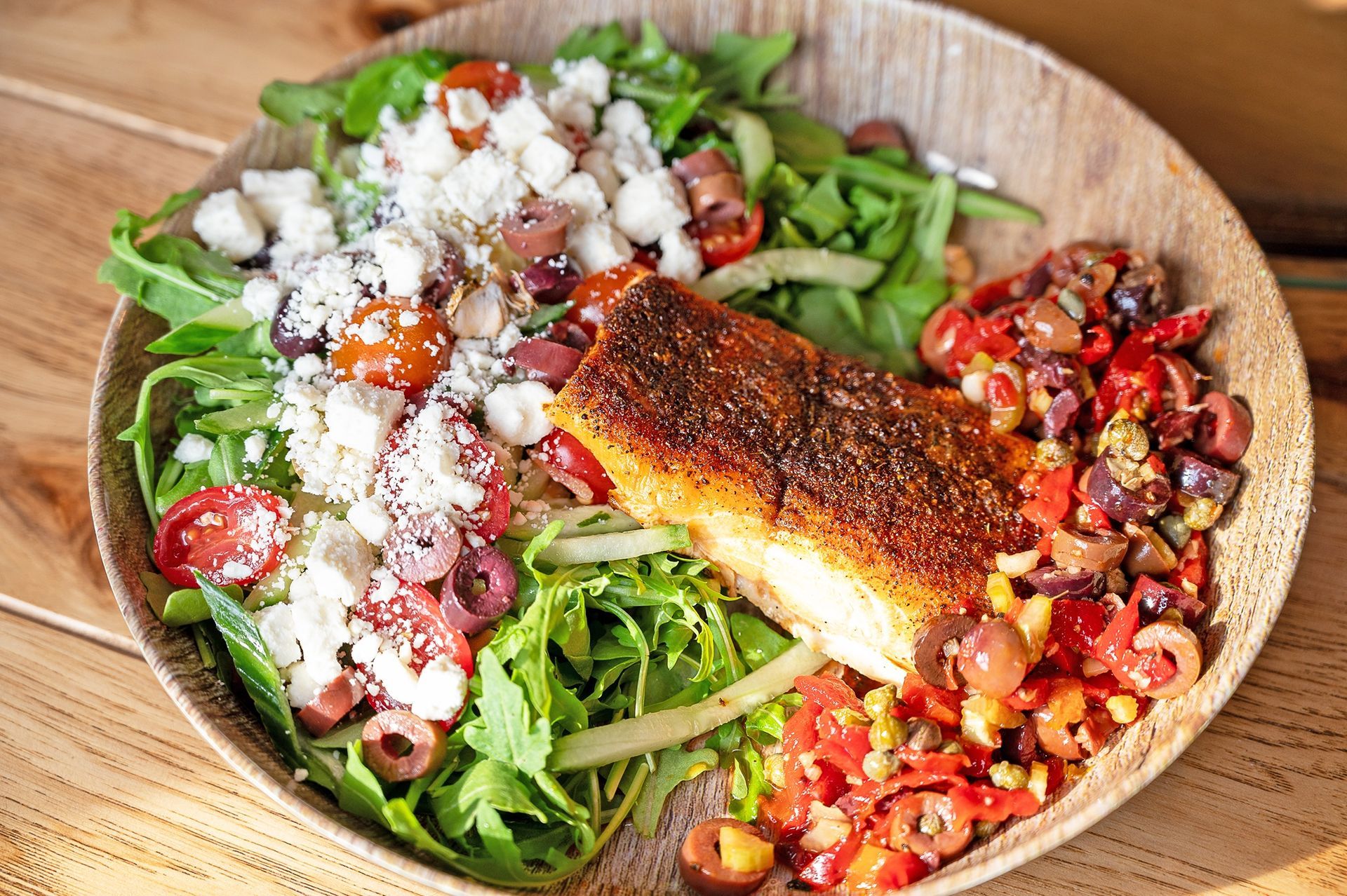 A bowl containing a seared fish fillet served over arugula, topped with feta, olives, and diced tomatoes on a wood table.