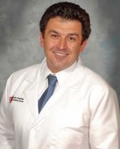 A smiling healthcare professional in a white coat and blue tie standing against a gray backdrop.