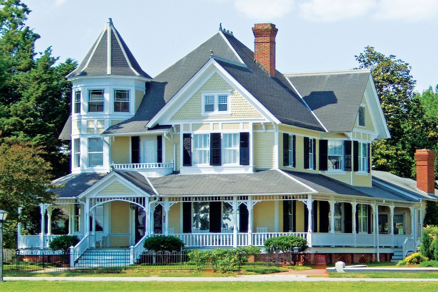 A two-story Victorian house with pale yellow siding, a wraparound porch, and a prominent turret, set among green trees.
