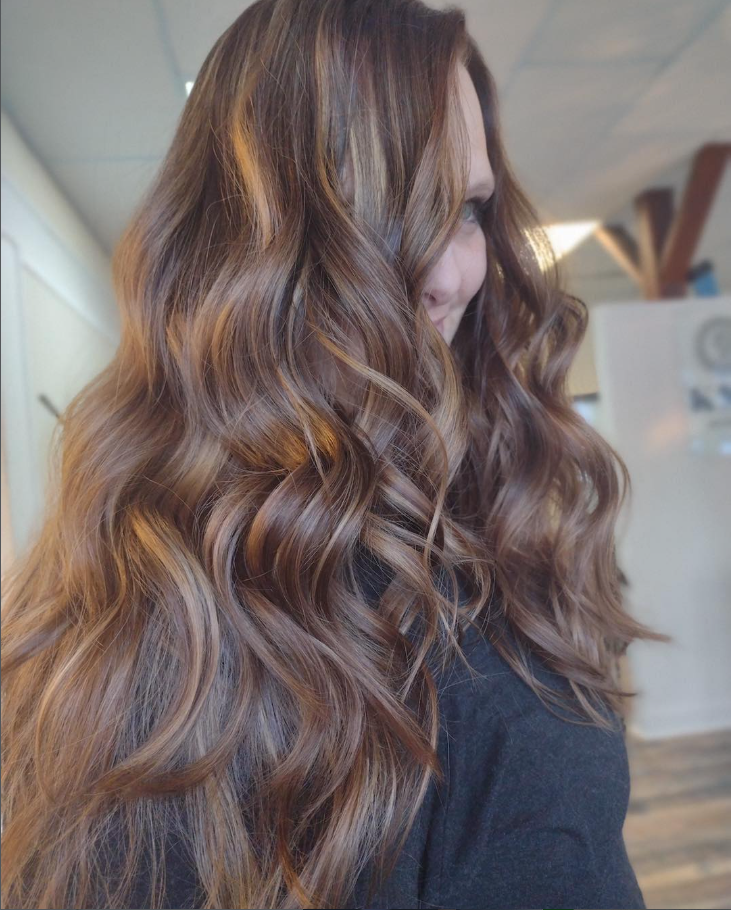 A person with long, wavy, chestnut brown hair featuring subtle caramel highlights, viewed from the side.
