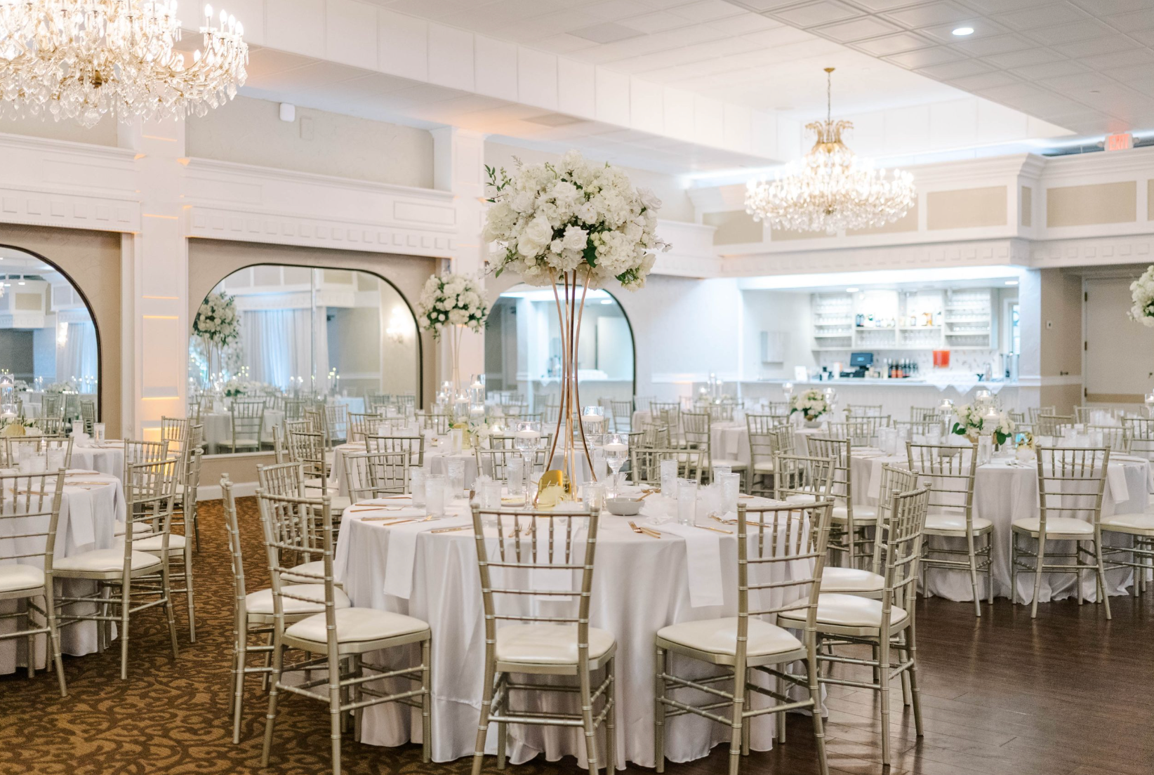 A bright, elegant banquet hall set for a formal event with round tables, white linens, and gold chairs under chandeliers.