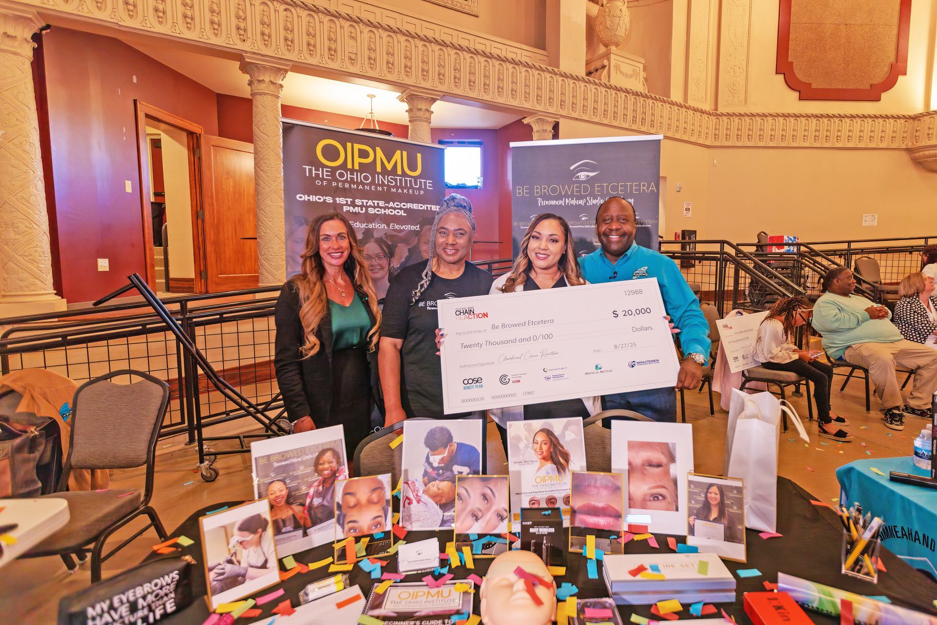 Four people hold a large ceremonial check behind a table covered with promotional materials and displays at an indoor event.