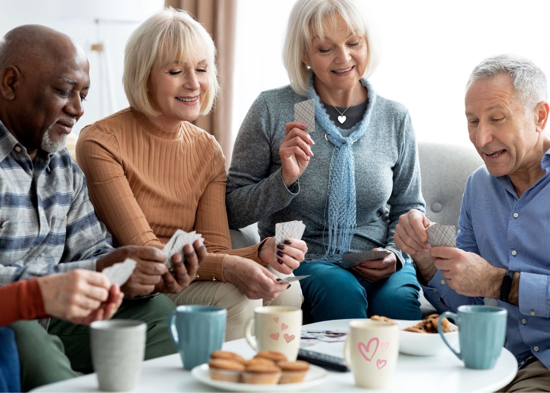 Four people sit around a coffee table, playing cards and drinking from mugs in a bright indoor setting.