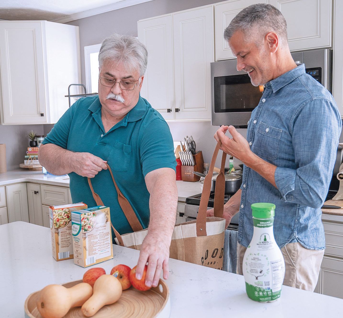 Two people in a kitchen unpacking groceries from paper bags onto a white counter, with produce and boxes visible.