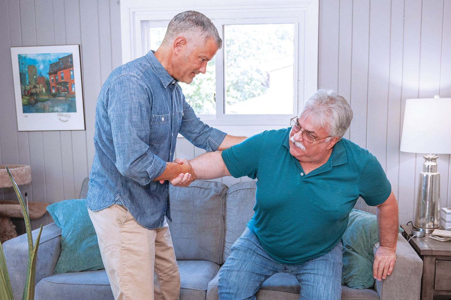 One person in a blue shirt holds the arm of another person in a green shirt to help them stand up from a sofa.