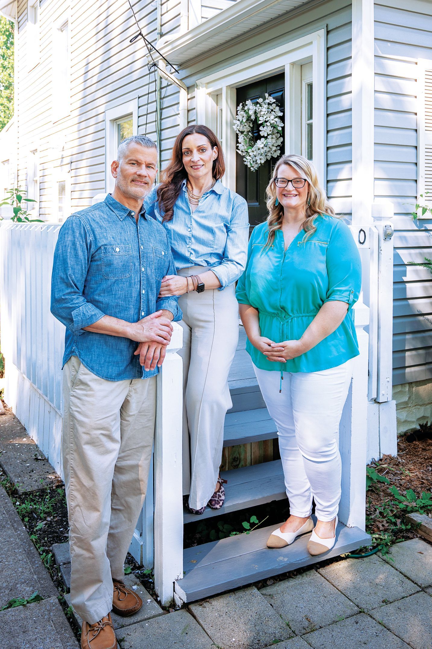 Three people pose on the porch steps of a light blue house, wearing casual clothing and smiling toward the camera.