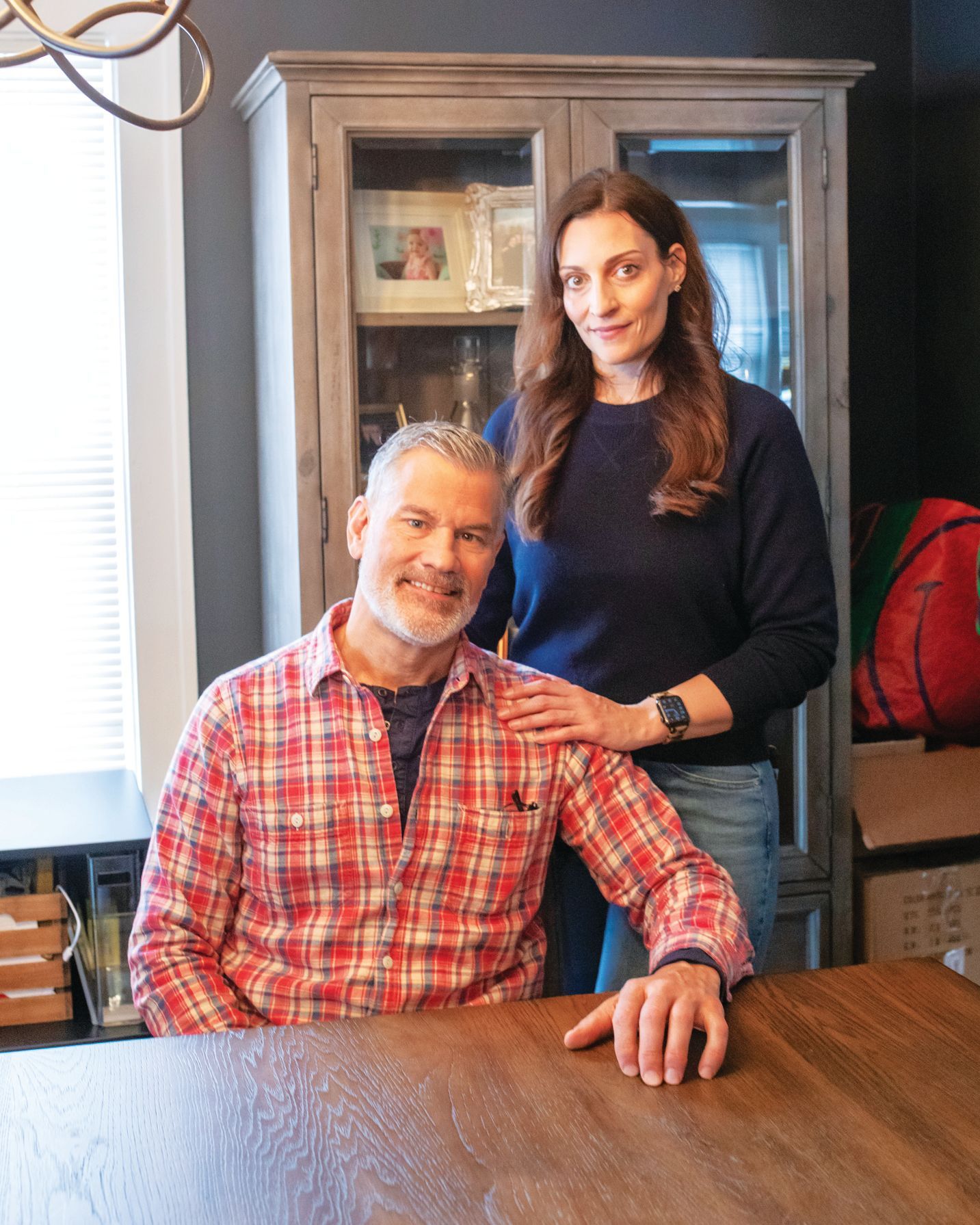 A person in a plaid shirt sits at a wooden table while another stands behind them, hand on their shoulder, in a room.