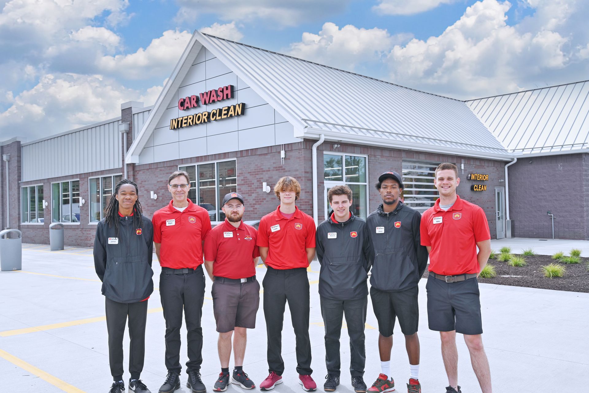 Seven team members in red shirts and uniforms stand together in front of a brick car wash building under a blue sky.