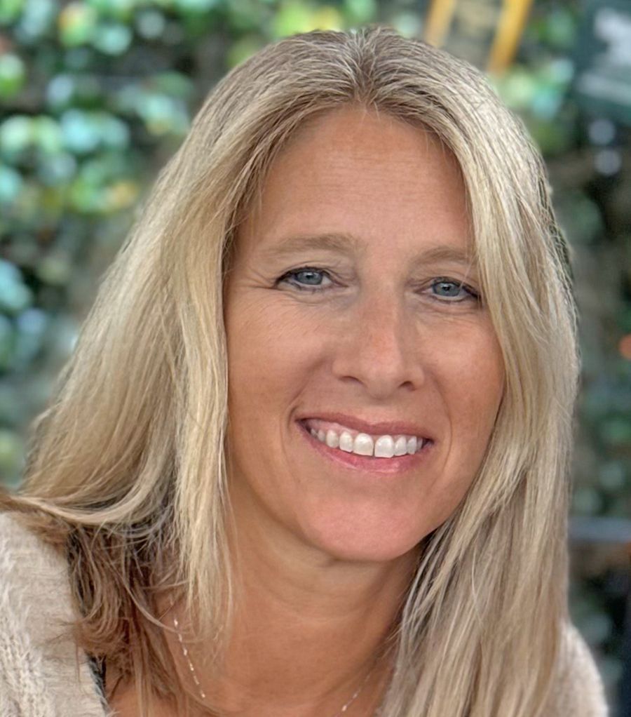 A close-up portrait of a smiling woman with long blonde hair, set against a blurred outdoor background.