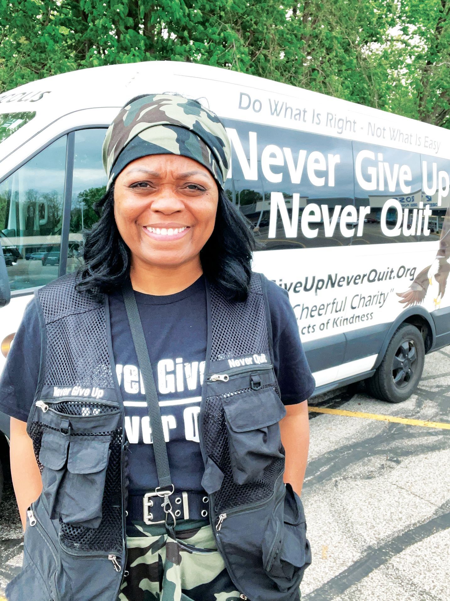 A smiling person wears a camouflage headscarf, black vest, and t-shirt in front of a white van with 