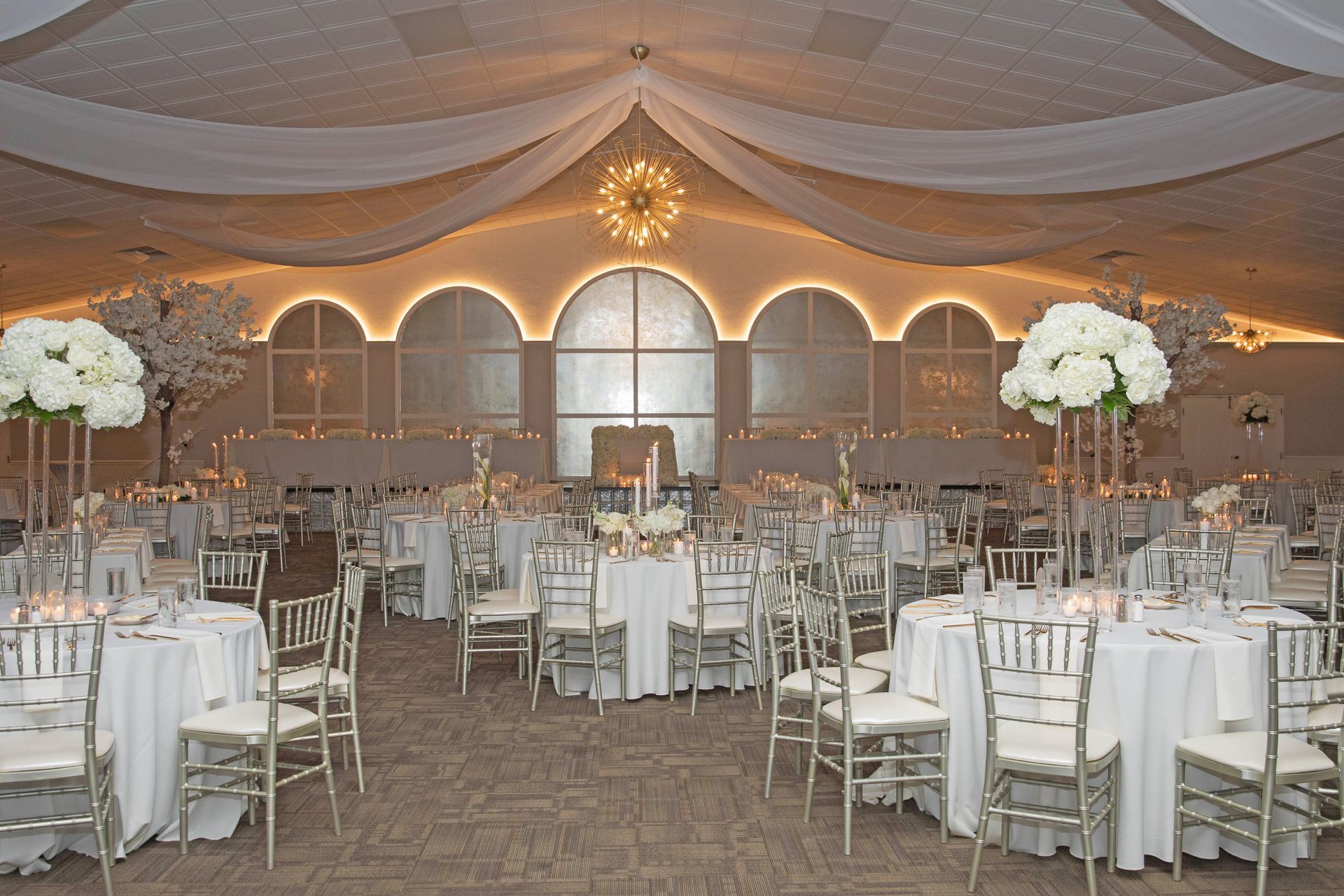 An elegant banquet hall set for a formal event with white round tables, silver chairs, and tall floral centerpieces.