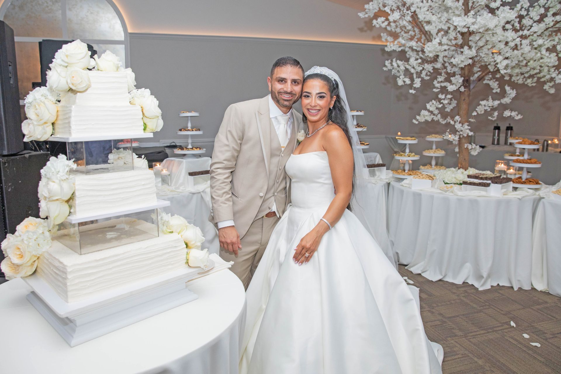 A smiling couple in wedding attire poses next to a tall, tiered white cake in a decorated event space.
