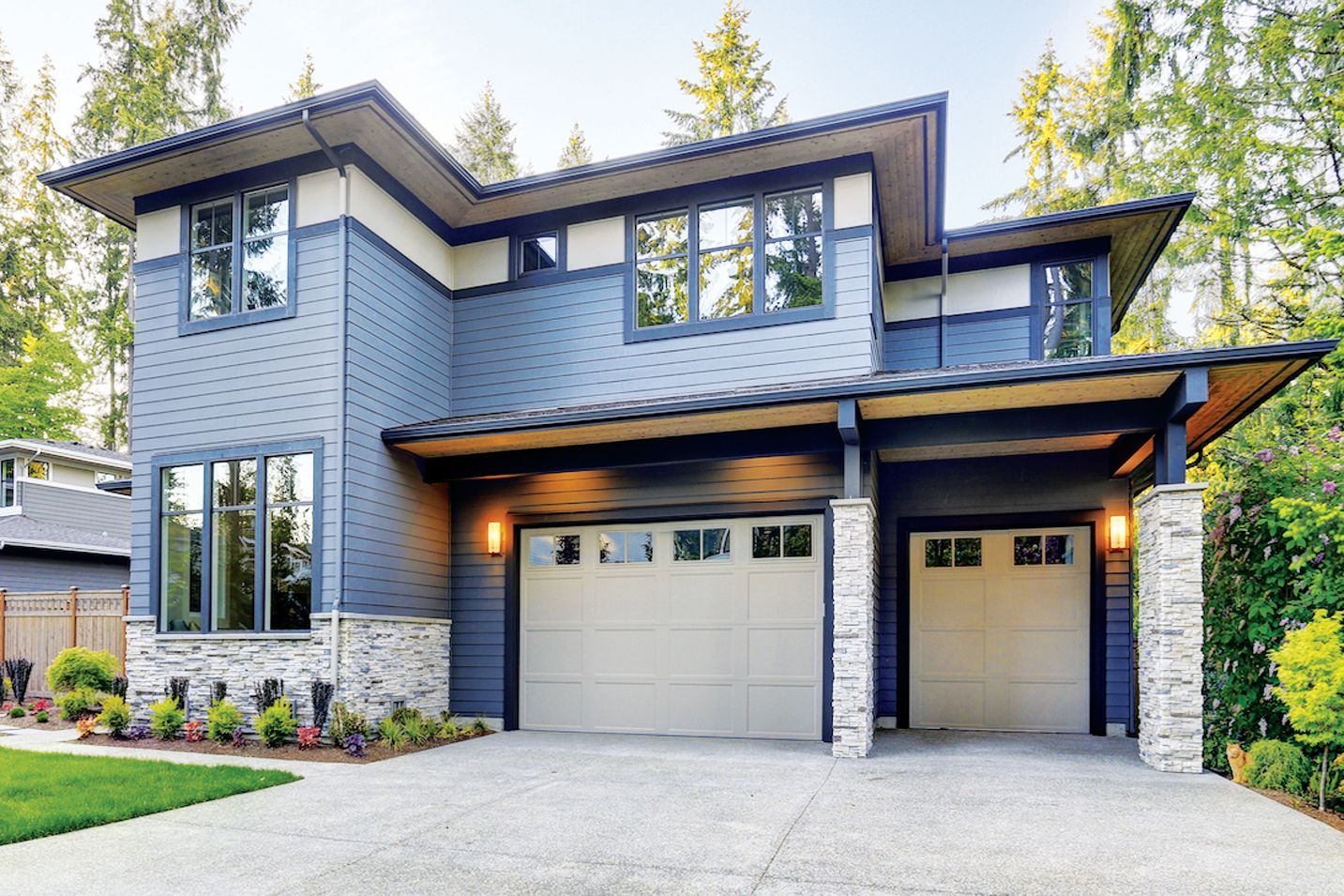 A two-story blue house with stone accents, a beige three-car garage, and a paved driveway, surrounded by trees.